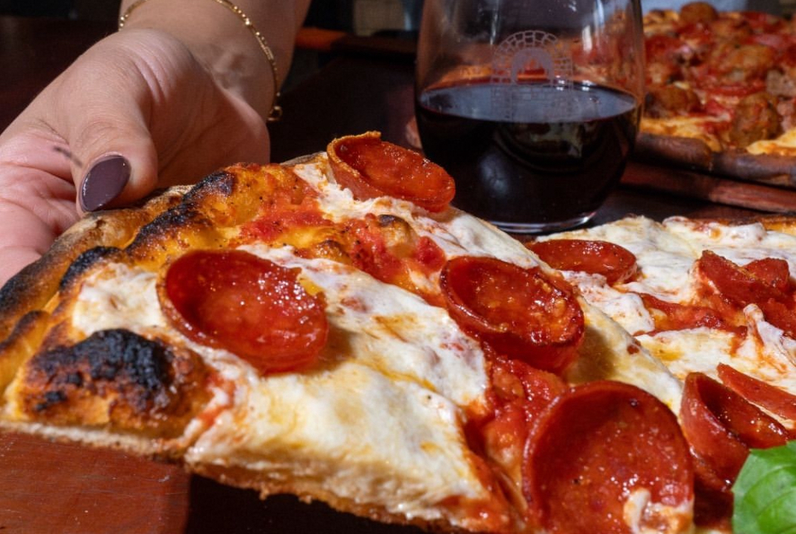 This photo shows a close-up of a slice of pizza topped with pepperoni and mozzarella, with a woman's hand removing the slice from a whole pie.