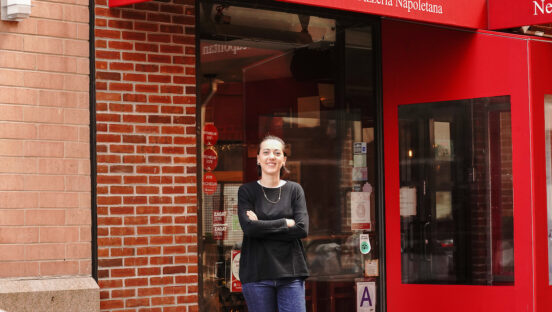 This photo shows a smiling Giorgia Caporuscio, wearing a black top and blue jeans, standing in front of Don Antonio with her arms crossed