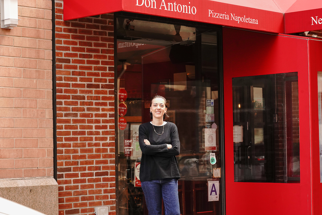 This photo shows a smiling Giorgia Caporuscio, wearing a black top and blue jeans, standing in front of Don Antonio with her arms crossed