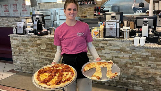 This photo shows a Fiori's Pizzeria server holding two trays of pizza, both designed with the Pi symbol.