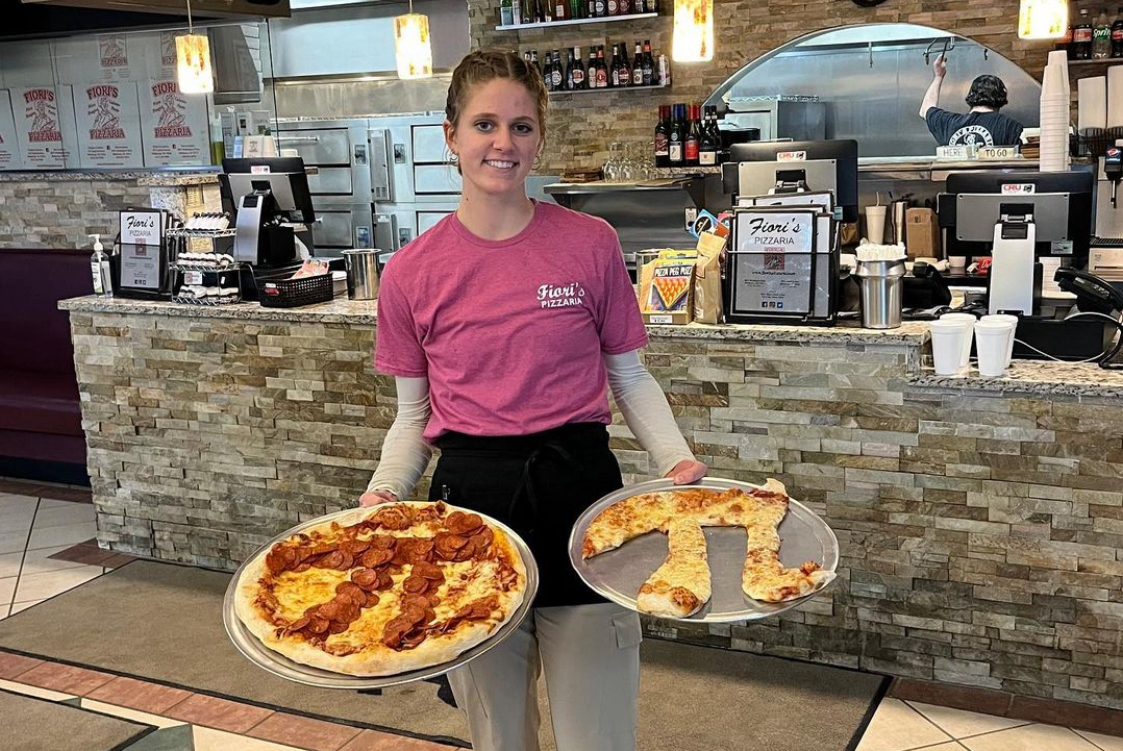 This photo shows a Fiori's Pizzeria server holding two trays of pizza, both designed with the Pi symbol.