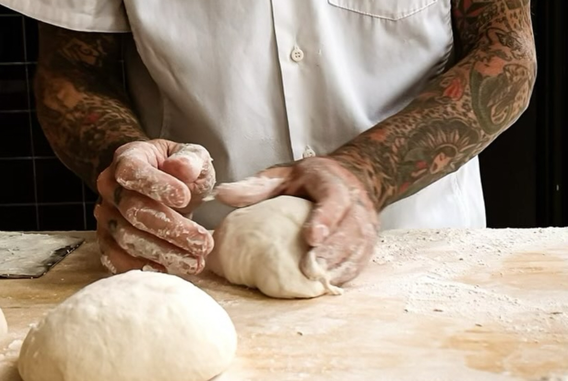 This photo shows Anthony Mangieri, shown from the chest down with heavily tattooed arms, kneading a ball of dough.