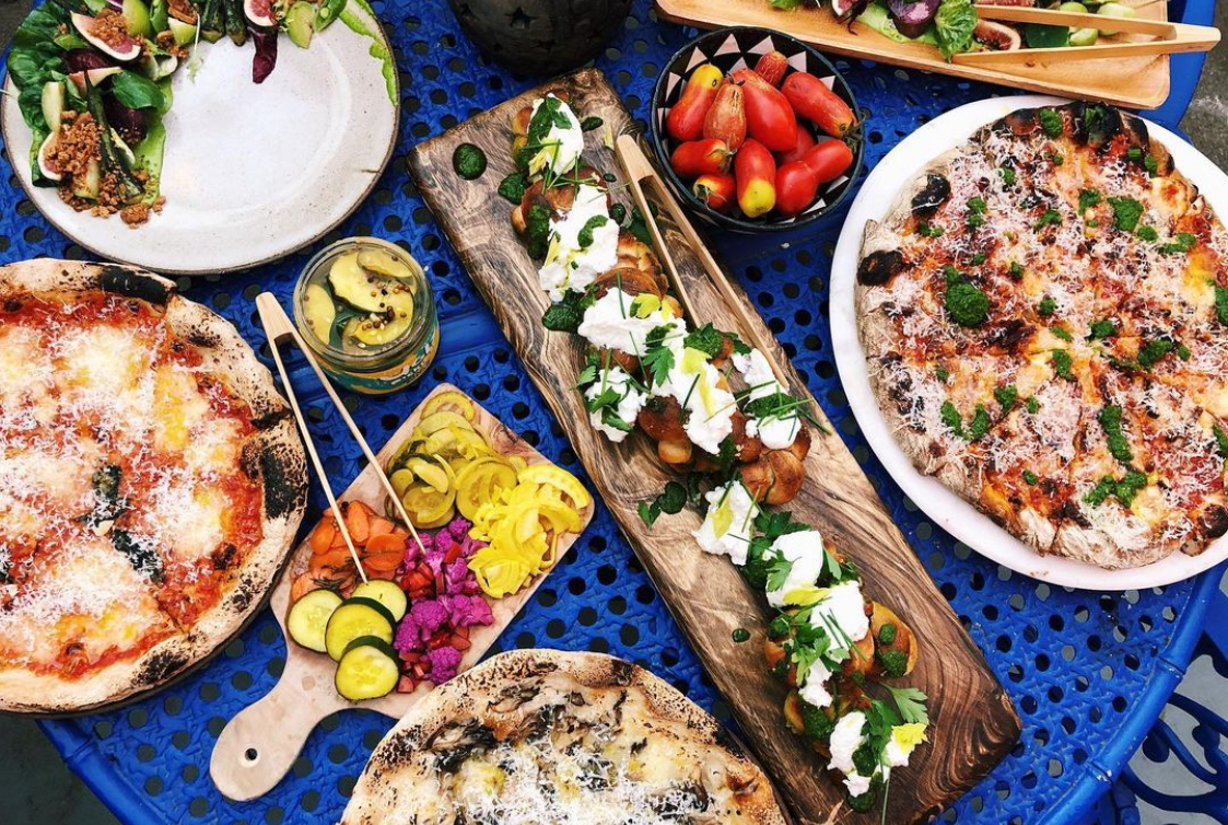 This photo shows a variety of menu items from Shuggie's, including pizzas and a salad, plus small plates of various imperfect vegetables.