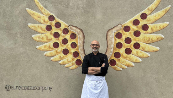 This photo shows Steve Bangos, arms crossed and wearing a black shirt and white apron, standing in front of his pizza wings mural.