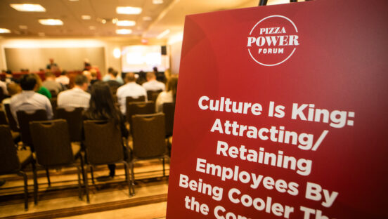 A picture of a sign at a conference, with the panel going on behind the sign.