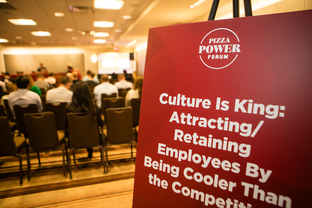 A picture of a sign at a conference, with the panel going on behind the sign.