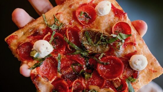 This photo shows a slice of a Grandma-style pizza topped with pepperoni, sausage, ricotta, bacon and more.