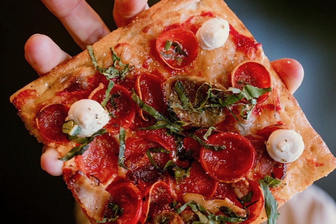 This photo shows a slice of a Grandma-style pizza topped with pepperoni, sausage, ricotta, bacon and more.