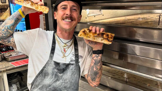 This photo shows Anthony Ludovici-DeBrigida, sporting a thick brown mustache, smiling broadly and holding up two slices of pizza to show off the air pockets in the crust.