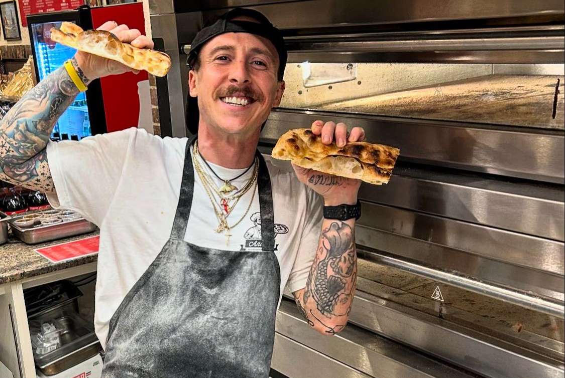 This photo shows Anthony Ludovici-DeBrigida, sporting a thick brown mustache, smiling broadly and holding up two slices of pizza to show off the air pockets in the crust.