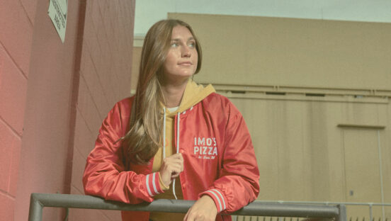 This photo shows a young woman with long brown hair wearing the red Satin Bomber Jacket from Imo's.