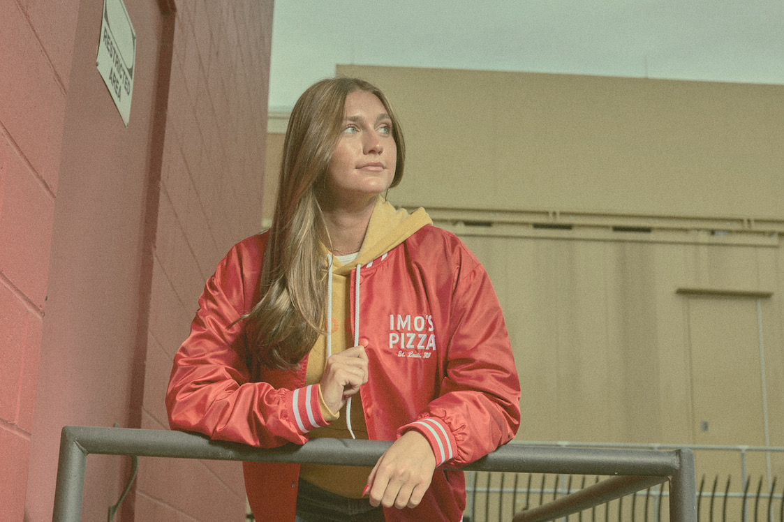 This photo shows a young woman with long brown hair wearing the red Satin Bomber Jacket from Imo's.