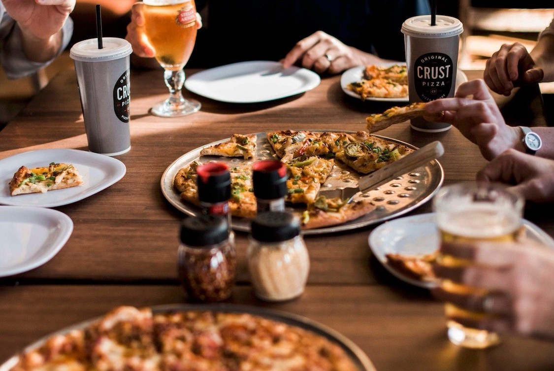This photo shows two customers with focus on pizzas, beer and a beverage cup at Crust Pizza Co.