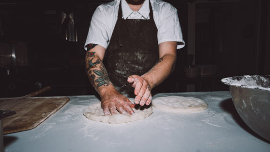 A pizzaiolo prepares a dough ball to be stretched. The shot is cut off from the neck down, so the pizzaiolo's face is not shown.