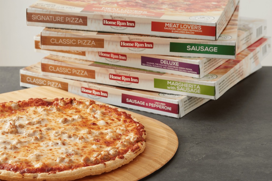 This photo shows a baked pizza with cheese and sausage placed in front of a stack of five Home Run Inn pizza boxes.