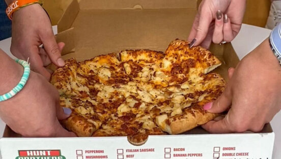 This photo shows several hands pulling out slices from a whole pizza in a Hunt Brothers-branded box.