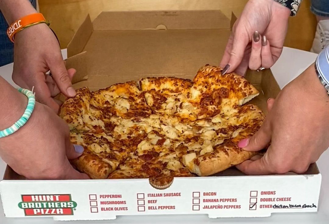 This photo shows several hands pulling out slices from a whole pizza in a Hunt Brothers-branded box.