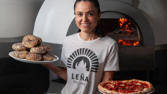 This photo shows Marisol Doyle, holding a plate of bagels in one hand and a pizza in the other, smiling and standing in front of her wood-fired oven.