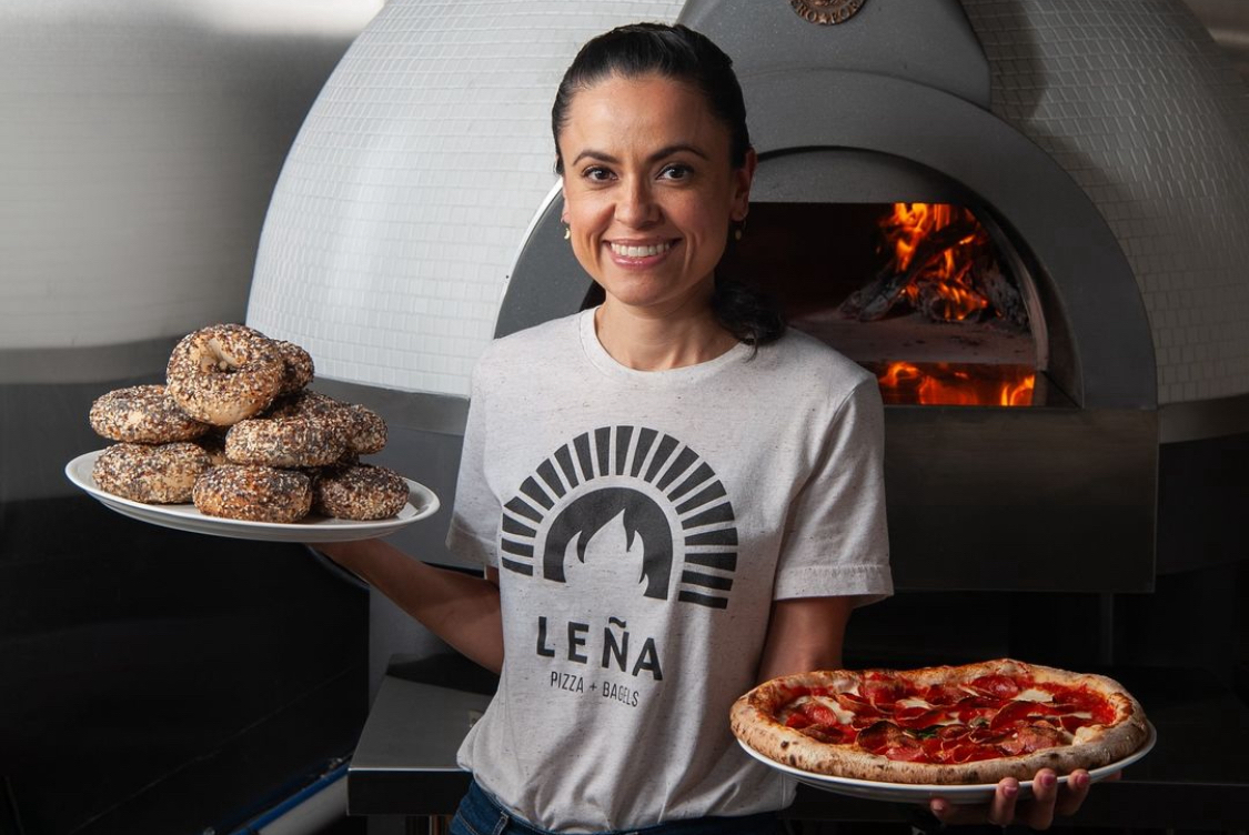 This photo shows Marisol Doyle, holding a plate of bagels in one hand and a pizza in the other, smiling and standing in front of her wood-fired oven.