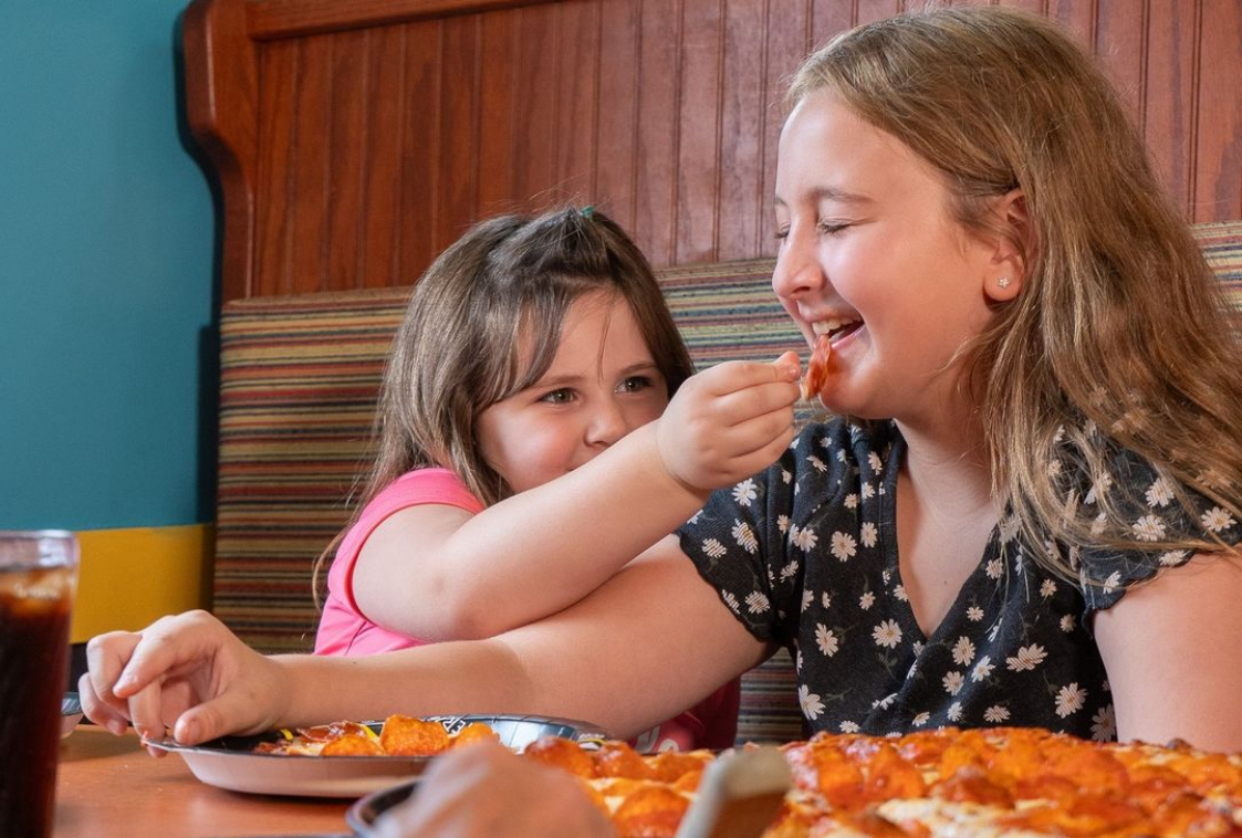 This photo shows a little girl putting a bite of pizza into an older girl's mouth while the older girl laughs.