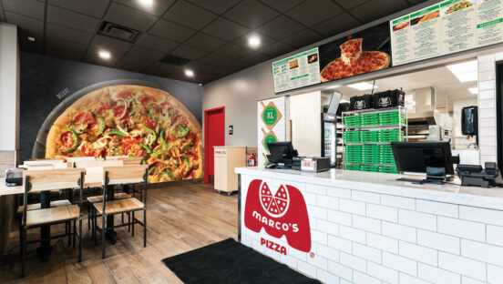 This photo shows the interior of a Marco's Pizza location, which includes a huge mural of a loaded pizza on a side wall and the ordering counter with the Marco's logo seen from the side.