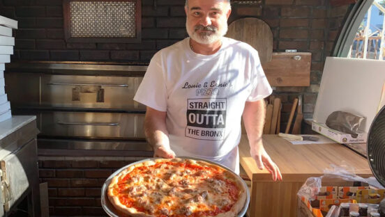 A man stands in a pizza kitchen with a whole cheese pizza on his peel.