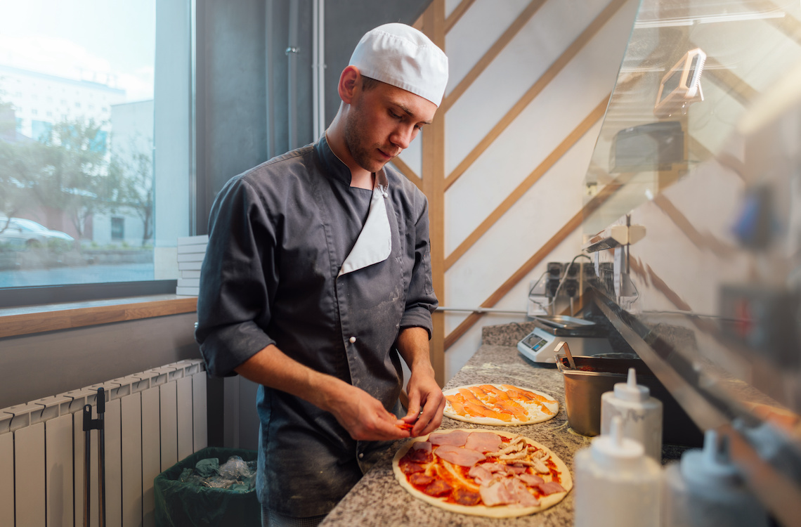 This photo shows a man wearing a dark gray chef jacket making a pizza in a kitchen.