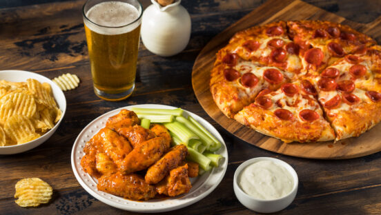 This photo shows a pepperoni pizza, chicken wings, a cup of dipping sauce, a glass of beer and a bowl of chips.