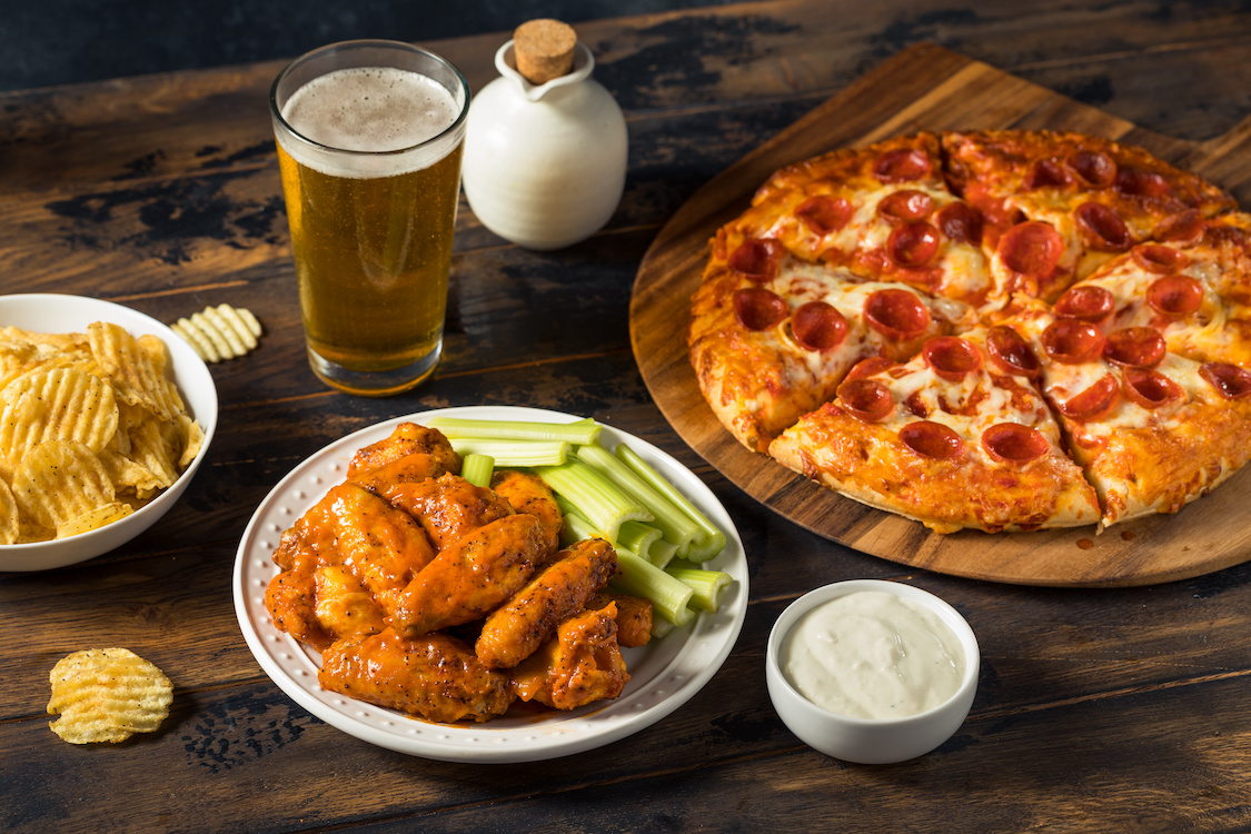 This photo shows a pepperoni pizza, chicken wings, a cup of dipping sauce, a glass of beer and a bowl of chips.