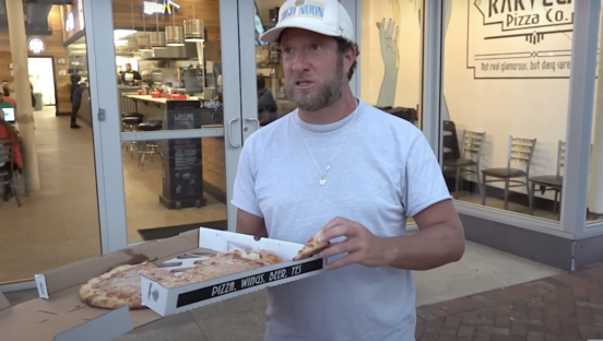 This photo shows Dave Portnoy, wearing a white t-shirt, standing out in front of Karvelas Pizza Co. with a box of cheese pizza.