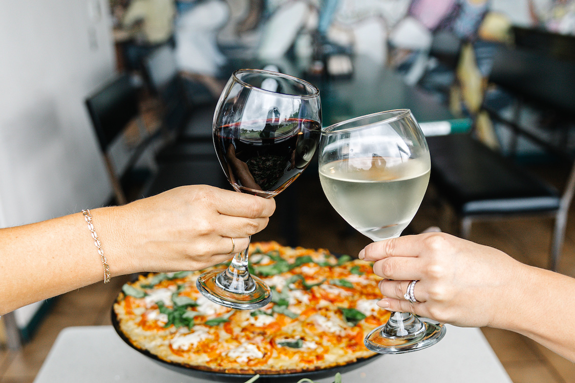 This photo shows two people clinking their large schooner glasses of wine together over a pizza.