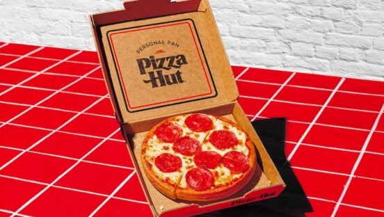 A personal Pizza Hut pan pizza in a box on red tiles with white brick in the background.