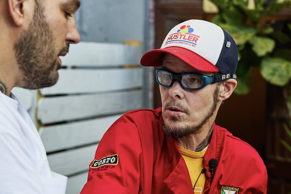 Auggie Russo, with a hat and Rex Specs, speaks to another pizzaiolo.
