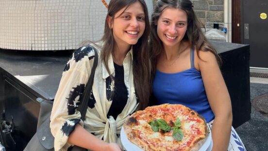 This photo shows two young women with long dark hair smiling and sitting shoulder to shoulder, with a Margherita pizza in their lap.