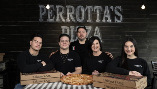 A family of Italian-Americans sits with a pizza in front of them and the backdrop on the wall reading "Perrotta's Pizza."