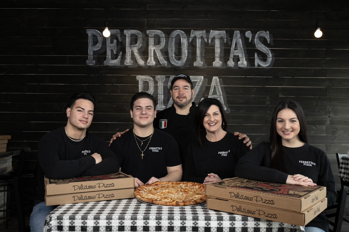 A family of Italian-Americans sits with a pizza in front of them and the backdrop on the wall reading "Perrotta's Pizza."