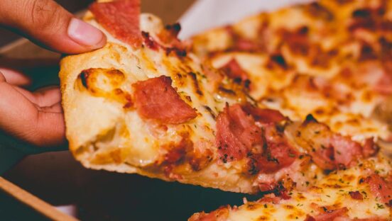This photo shows a person's hand removing a slice of pizza from a whole pie topped with mozzarella, pepperoni and Canadian bacon.
