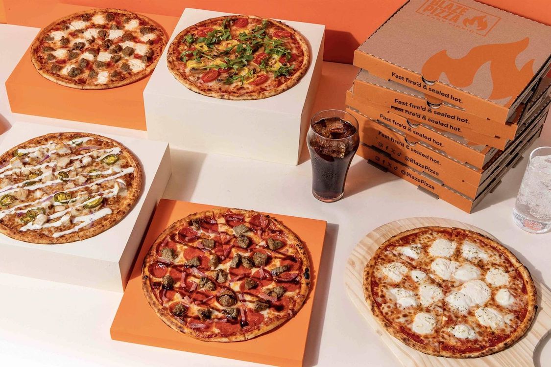 This photo shows a variety of pizzas along with a glass of iced cola and a stack of three Blaze Pizza delivery boxes.