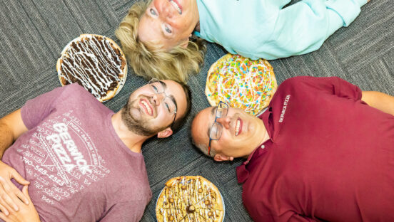 This photo shows Austin, Todd and Brenda Buckland lying on the ground, smiling and looking up at the sky, with several dessert pizzas next to their heads.