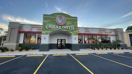 This photo shows the exterior of a Chuck E. Cheese restaurant, with the parking lot in front and a blue sky in the background.