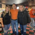 This photo shows Matt Plapp with the owners of Capuano's Pizza House inside their restaurant. He is standing between the two women with an arm around both, and they're all smiling happily.