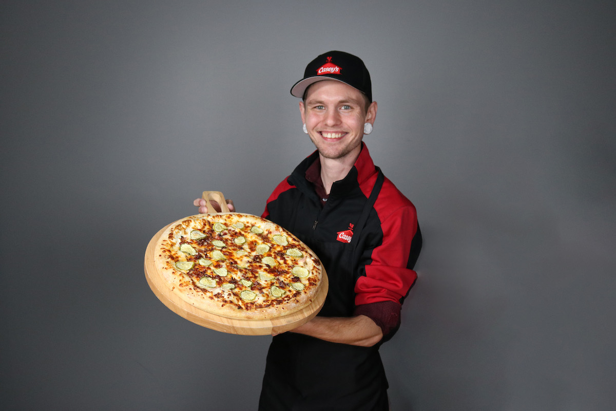 A chef in Casey's gear holds a pickle pizza.