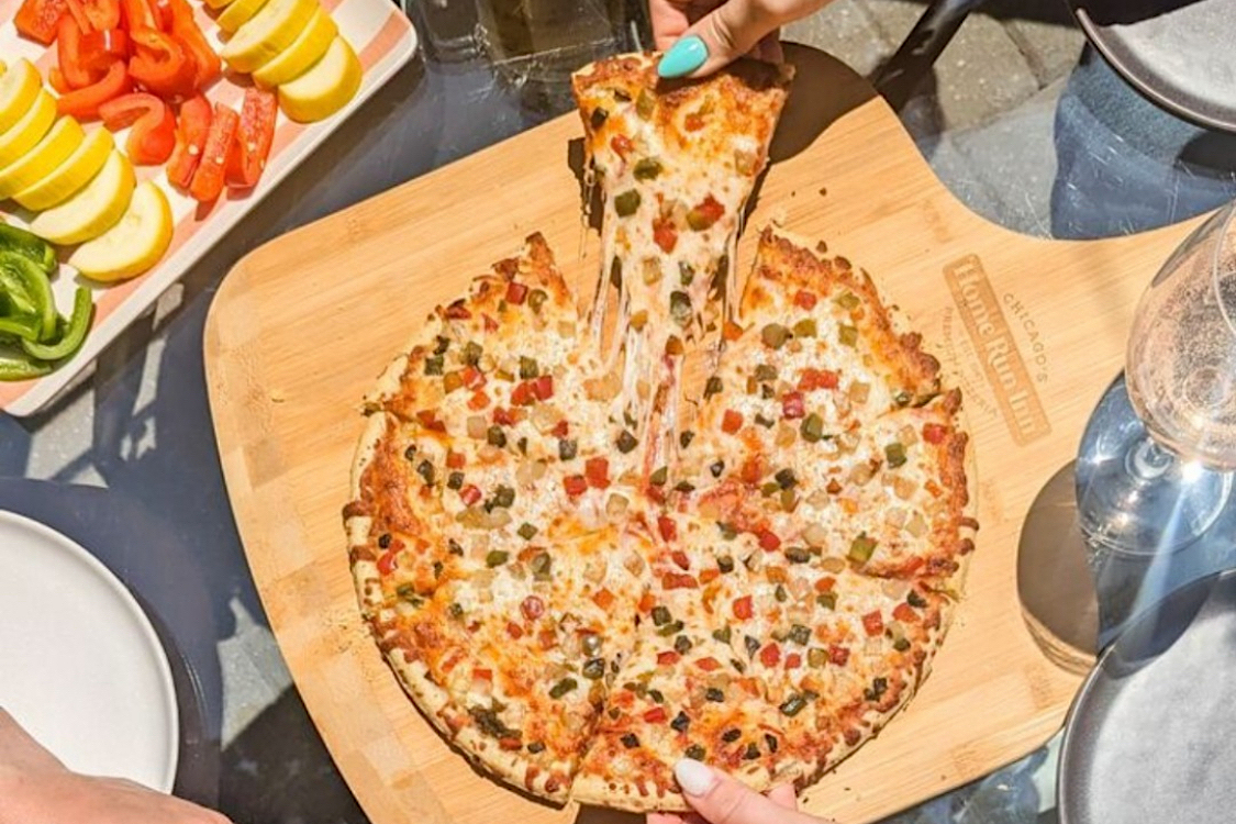 This photo shows a woman's hand, with fingernails painted a light blue, pulling a slice from a whole pie topped with sausage and peppers.