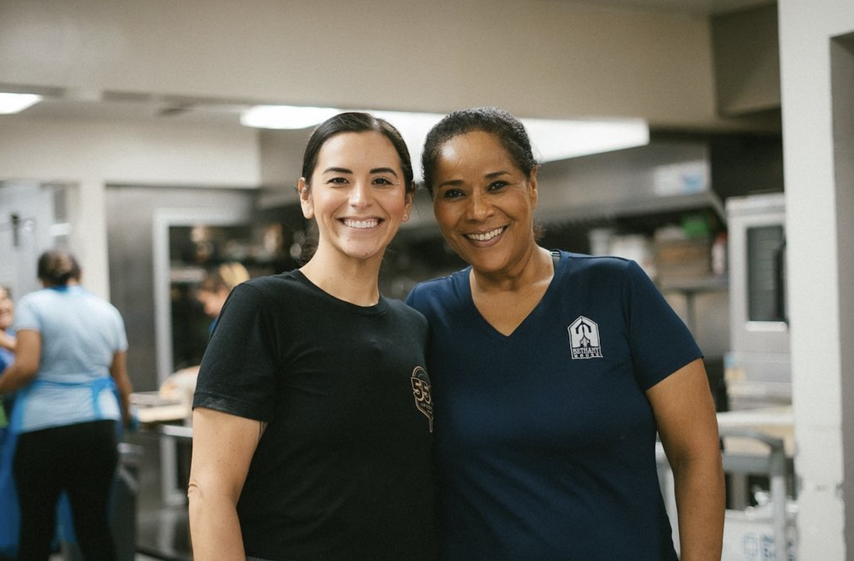 Two women stand, smiling at the camera, in a restaurant kitchen.
