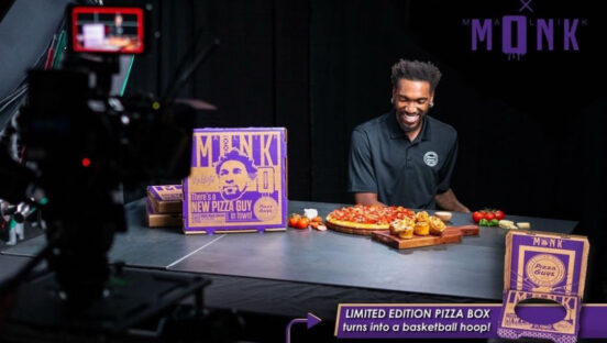 This photo shows Malik Monk, wearing black, shooting a video for Pizza Guys, with a special limited-time pizza box and a four-meats pizza sitting on a table in front of him. He is smiling and looking down.