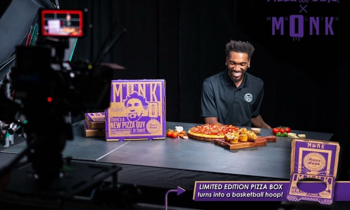 This photo shows Malik Monk, wearing black, shooting a video for Pizza Guys, with a special limited-time pizza box and a four-meats pizza sitting on a table in front of him. He is smiling and looking down.