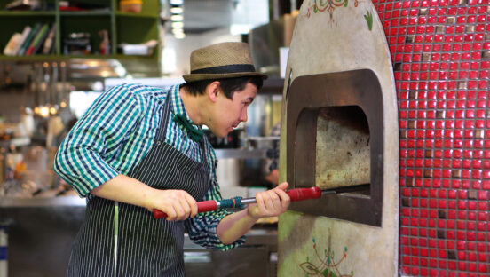 This photo shows a dark-haired man, wearing a blue and black striped shirt, black apron and an old-fashioned brown hat, peering into a wood-fired pizza oven while holding a peel.