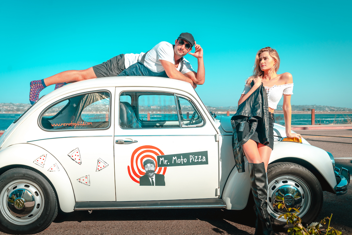 This photo shows a man, wearing sunglasses and a cap, draped over the roof of a white Volkswagen (branded with the Mr. Moto logo) and an attractive young woman in knee-high black leather boots leaning against the side of the car.