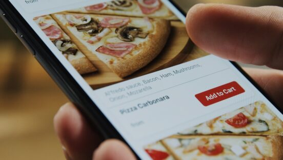 This photo shows a close-up of a pizzeria's ordering system on a phone's screen as a person's thumb hovers above a picture of a pizza slice.