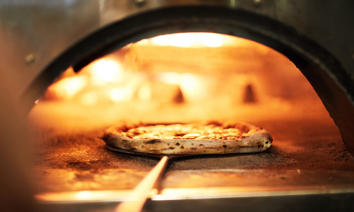 This photo shows a pizza with a beautifully blistered crust being peeled out of a wood-fired oven.